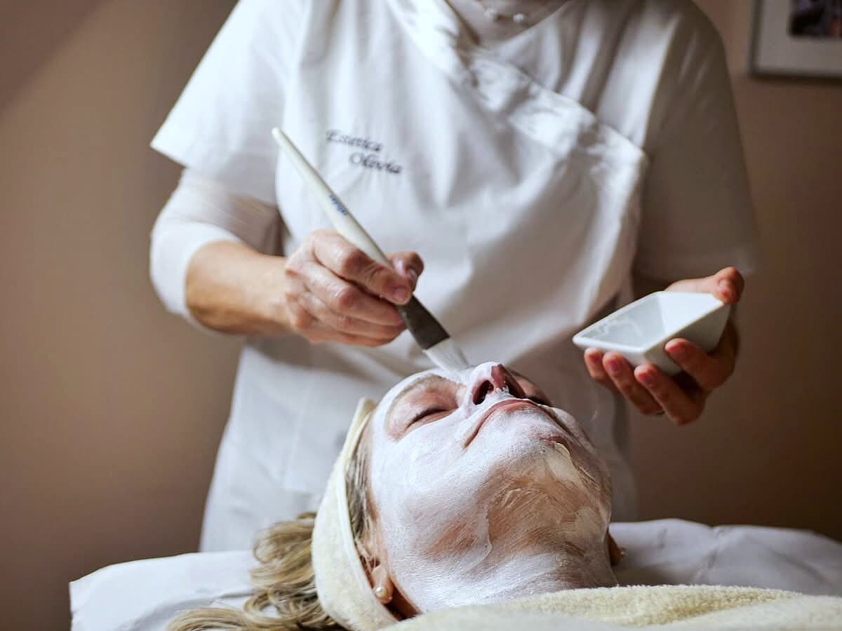 L'estetista con il camice ricamato Estetica Olivia stende la maschera sul viso di una cliente
