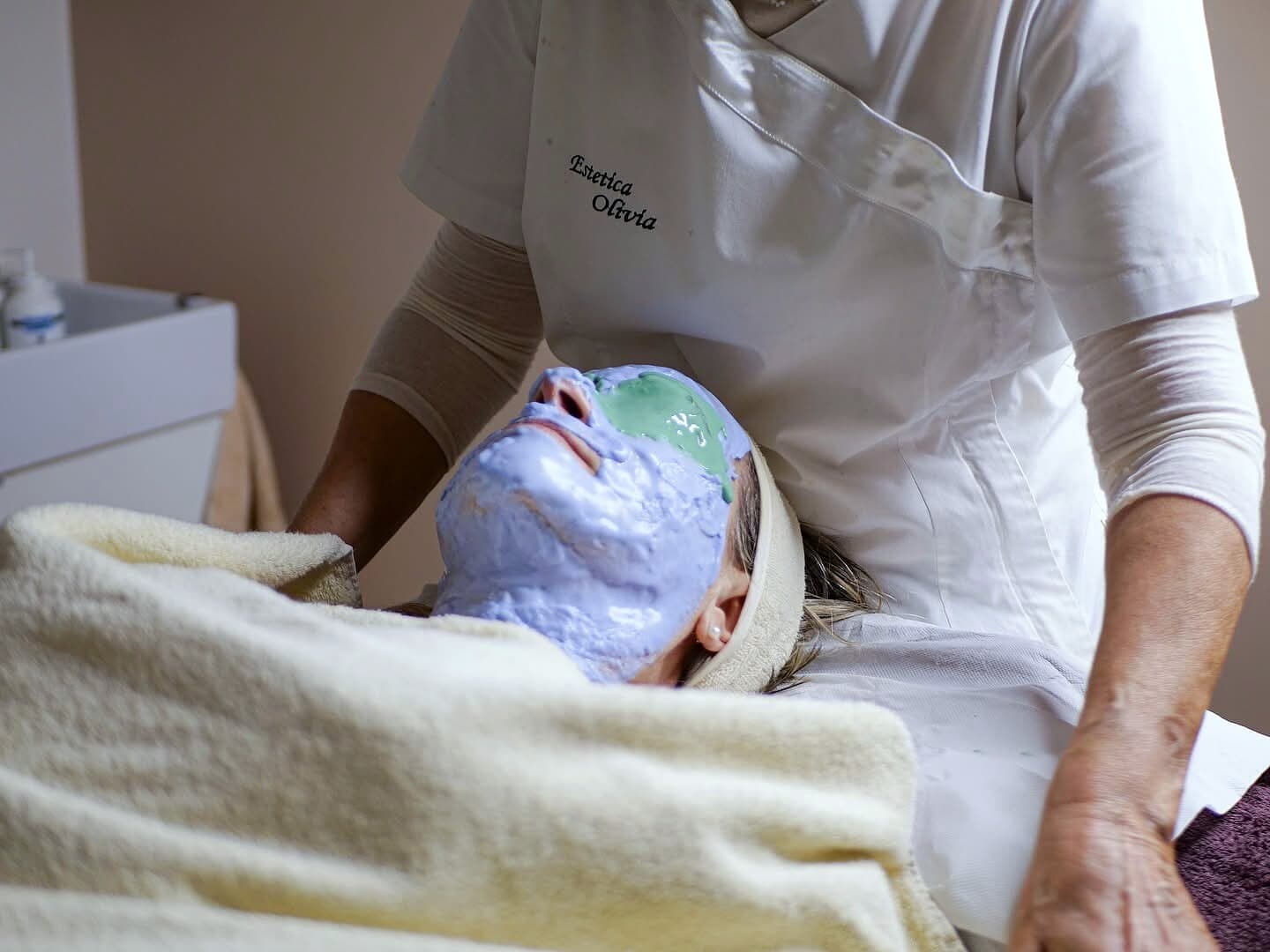 Una cliente distesa con la maschera lilla e verde sul viso, accanto il camice ricamato Estetica Olivia
